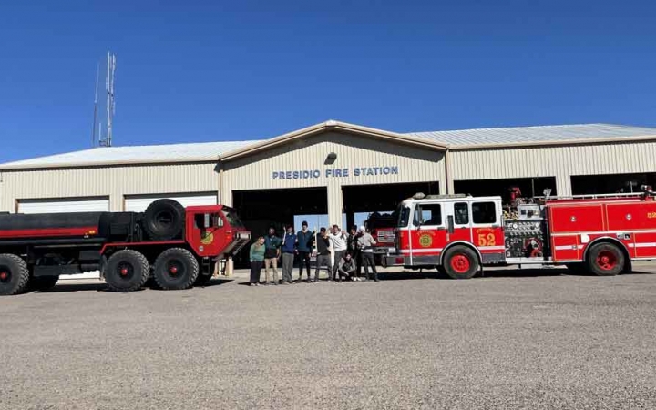 a group of students stand in front of a fire station and fire trucks while participating in a service project with outward bound